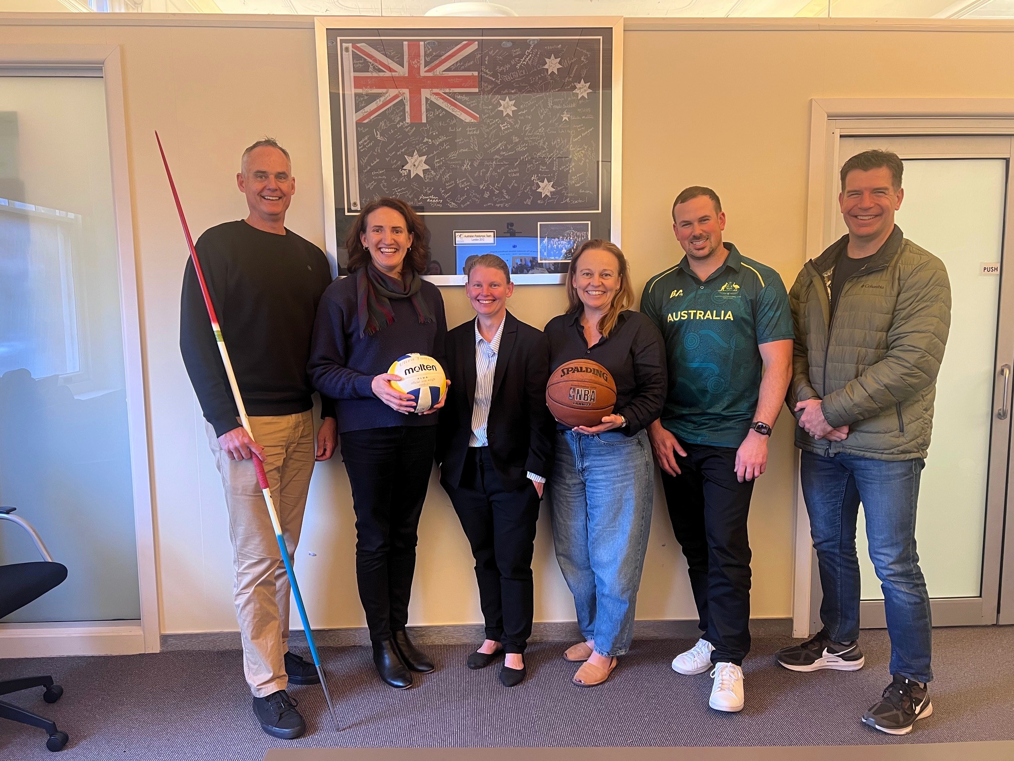 A group of six people are standing indoors in front of a bulletin board, posing together. One individual is holding a javelin, another is holding a volleyball, and another is holding a basketball. The group is dressed in a mix of casual and sports-themed clothing, including an Australia sports shirt, and they appear to be in an office or meeting space.