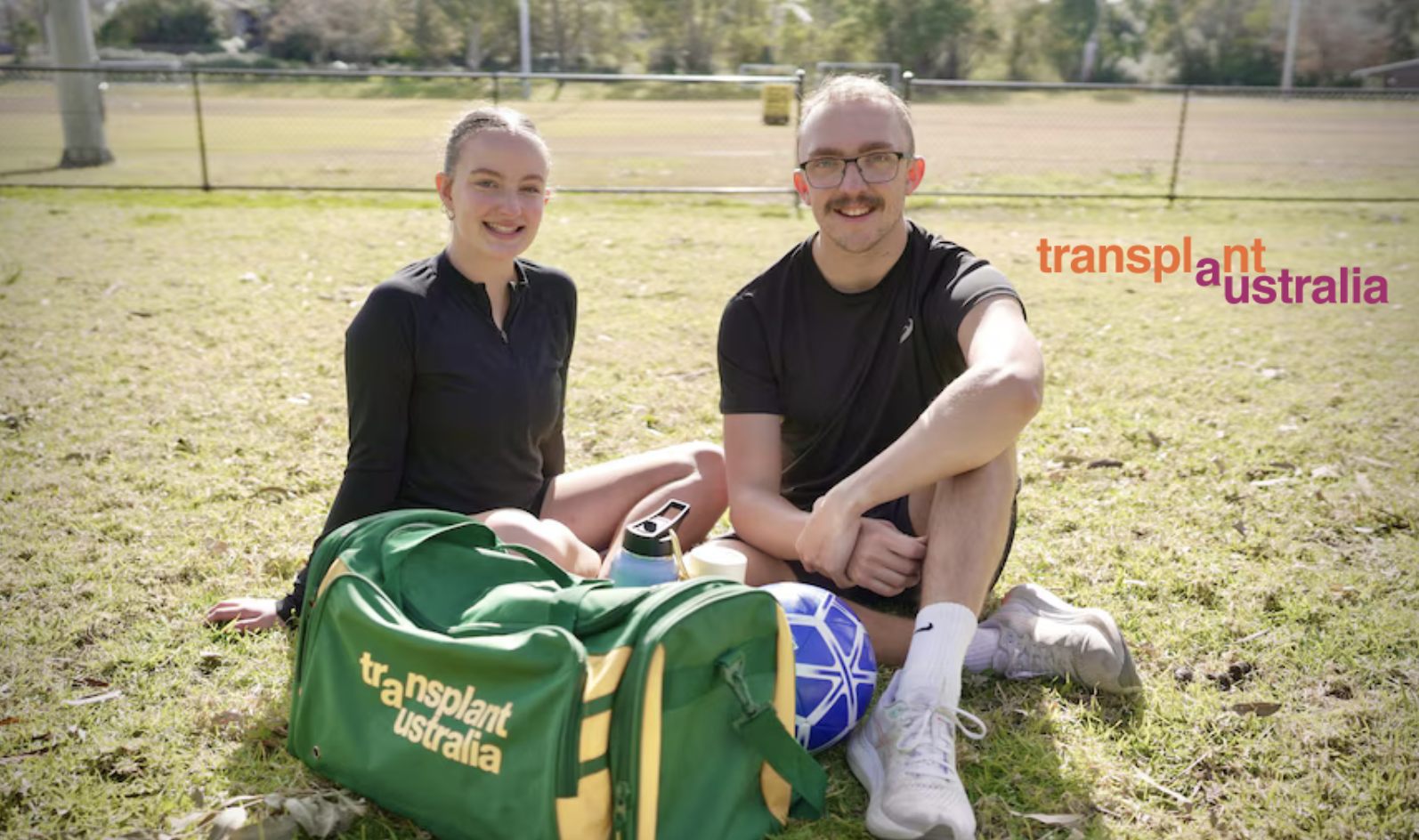 Two siblings in black athletic clothes are sitting on the grass in a sunlit outdoor field, with a blue soccer ball and a green and yellow sports bag labeled "transplant australia" in front of them. The Transplant Australia logo appears in orange and purple text on the right side of the image.