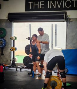 Inside a gym, a person in a weightlifting suit stands beside a bench with a blue water bottle; another person stands behind offering support by resting hands on their shoulders. A third individual bends over in front of a barbell with yellow "ROGUE" brand weight plates. Weight racks and equipment line the wall behind them, and the scene is illuminated by natural light from large windows. A sign above reads "THE INVICT..." (partially visible).
