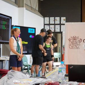 Four people are present in a gym or event setting. One person stands in front wearing weightlifting gear and appears to be the focus, supported by two others beside and behind them. A woman stands to the left, watching. In the background, screens show competition details, including the name "Eli Frew" and "48 kg." There are chairs, certificates on the wall, and foreground table items like water bottles and clothing.