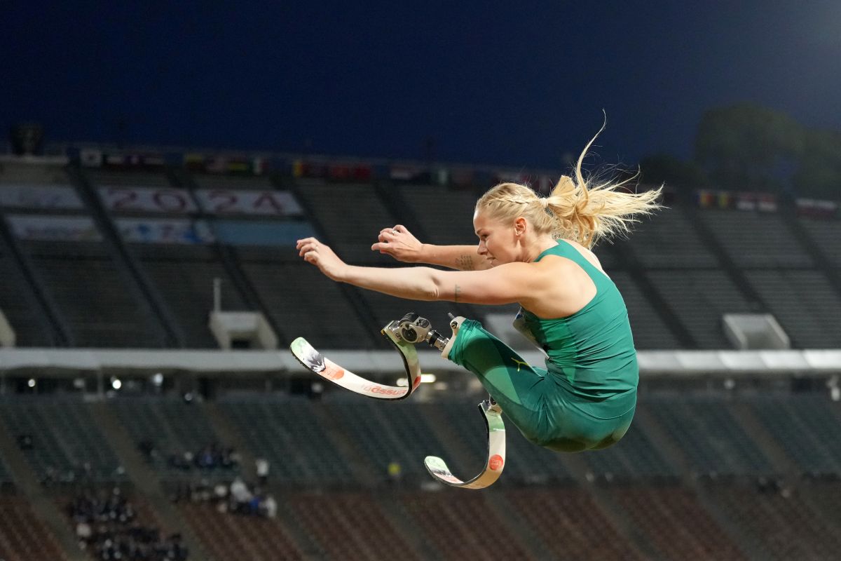 Image shows a Paralympic athlete with a prosthetic leg jumping in the air. She has a green jumpsuit on and blonde hair.