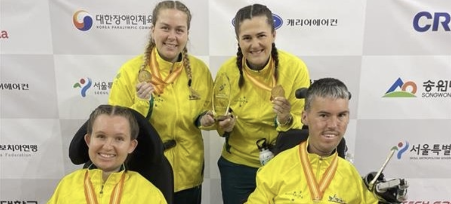 Image shows Boccia stars Dan Michel and Jamieson Leeson, and their respective ramp assistants Ash Maddern and Jasmin Haydon in front of a grey and white sign. They are all holding up gold meals and all wearing yellow jackets.