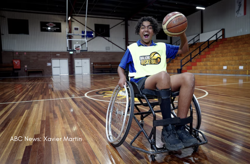 Image shows Quade Stone sitting in a wheelchair in an empty gymnasium. He is holding a basketball in one hand is making an expressive, happy face.