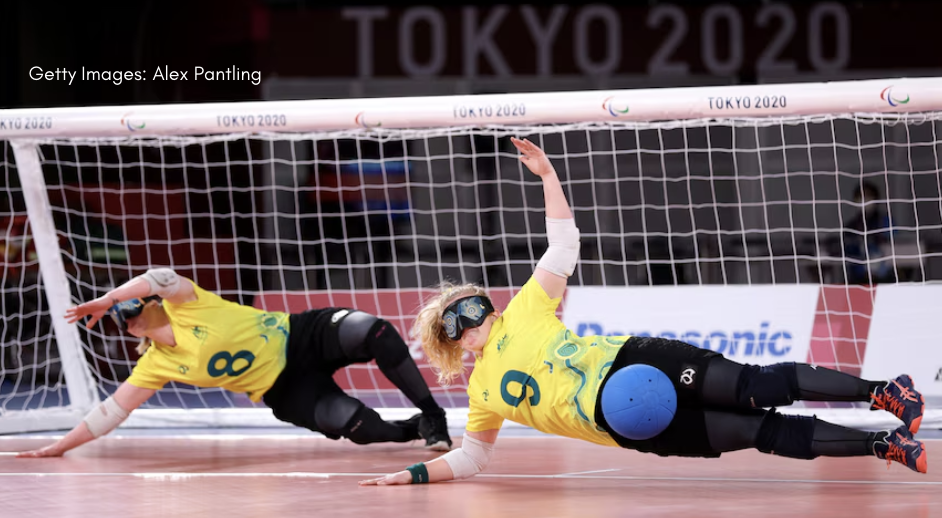 Image shows two goal ball players in action. They are both wearing Australian jerseys with black pants. They have eye masks on, and they are stopping a blue ball from going in the goal.