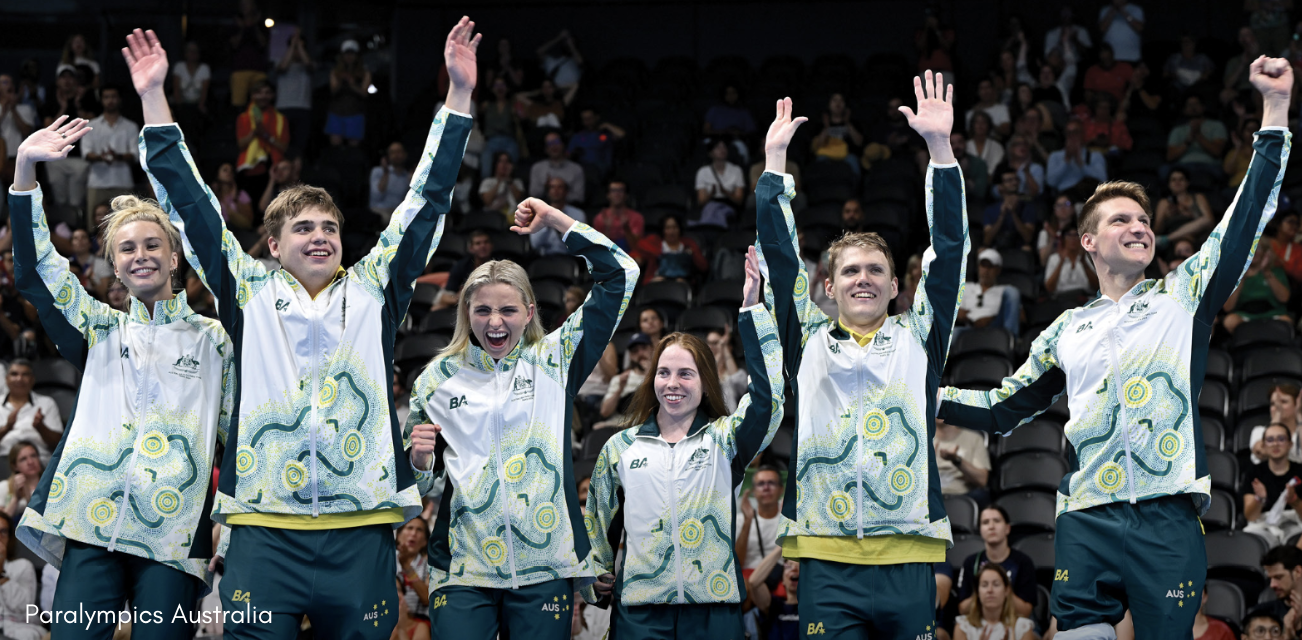 Image shows 6 athletes all standing with their hands in the air. They are all wearing green and gold paralympic uniforms.