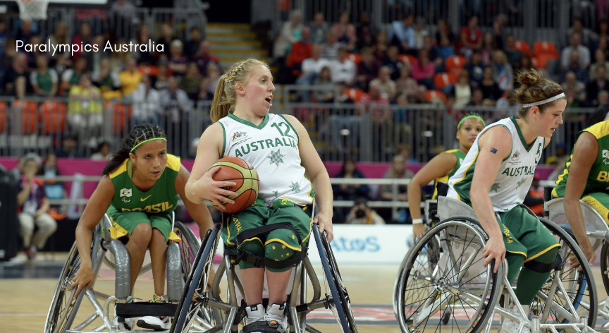 Australian and Brazilian women's wheelchair basketball players competing on court, with one Australian player pushing her wheelchair while holding a basketball. The arena is full of spectators and the players are wearing their respective team uniforms. The Paralympics Australia logo is visible in the top corner.