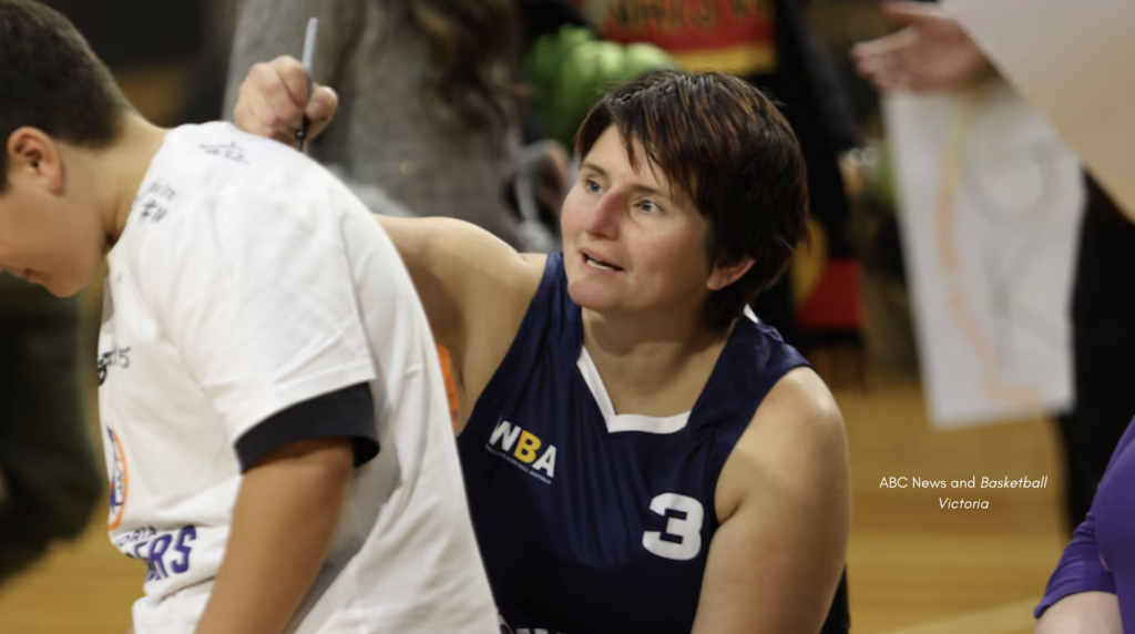 A wheelchair basketball player in a navy blue jersey with the number 3 signs the back of a child's white shirt at a basketball event. The scene is indoors with people and sports banners visible in the background. Image credit: ABC News and Basketball Victoria.