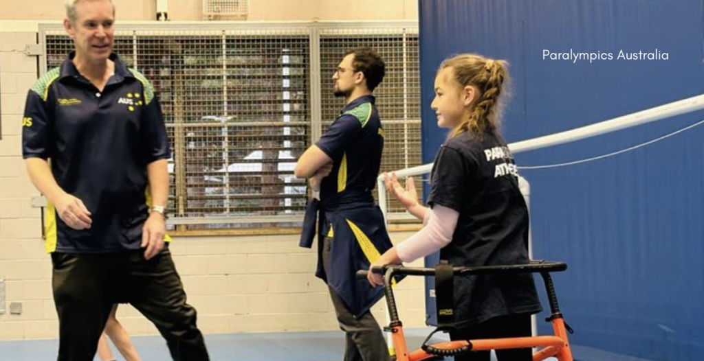 A coach and a young girl participate in an indoor sports session. The girl, wearing a "Paralympic Athlete" shirt and using an orange walker, faces the coach on a blue court. Another man stands in the background, watching the activity. The image is branded with "Paralympics Australia" in the top right corner.