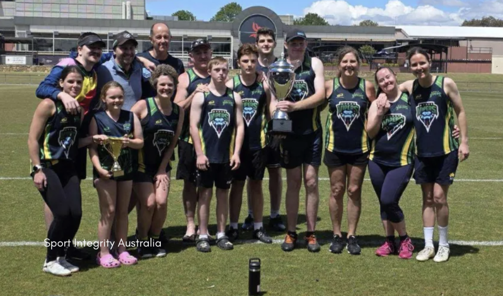 A group of athletes and supporters pose together on a grass sports field, smiling and holding two trophies. They are wearing matching sleeveless shirts with "Vipers" team logos and shorts. The background features a large building and blue sky. The image includes the watermark "Sport Integrity Australia" in the bottom left corner.