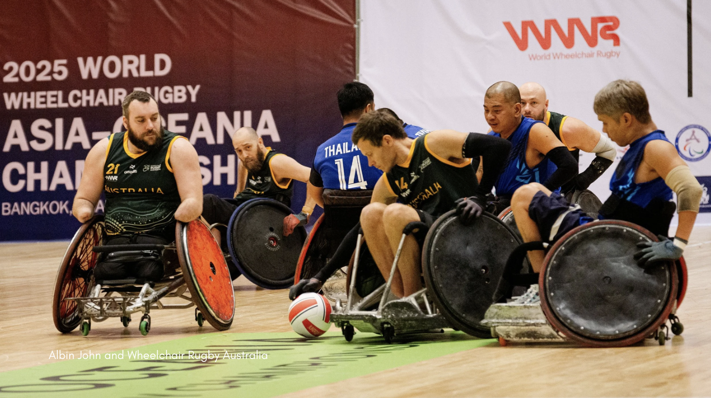 Australian and Thai wheelchair rugby players compete during the 2025 World Wheelchair Rugby Asia-Oceania Championship in Bangkok, with Australian athletes in green and gold jerseys challenging Thai players in blue as a player in an Australian chair controls the ball on court, and event branding visible on the backdrop.