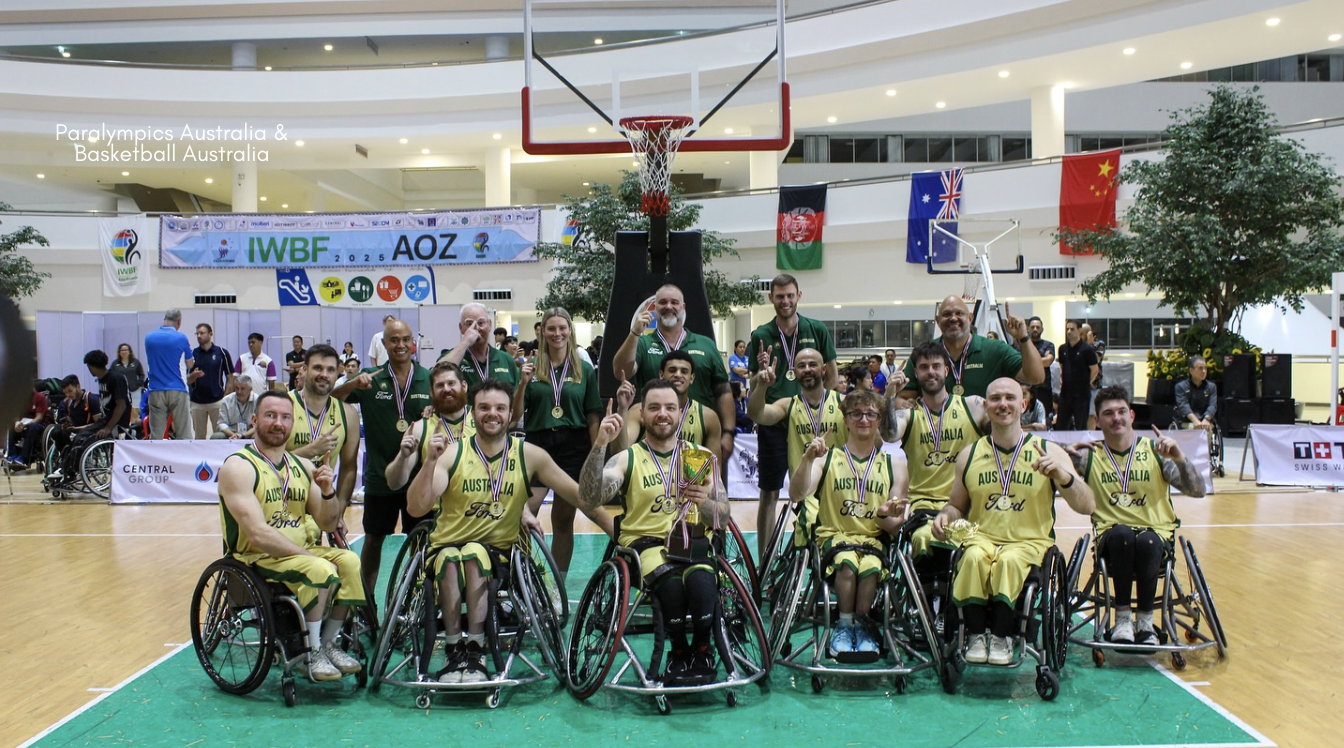 The Australian Rollers men’s wheelchair basketball team, wearing gold and green uniforms, celebrate on court in front of a basketball hoop in Bangkok after winning the 2025 IWBF Asia-Oceania Championships, proudly displaying gold medals and holding up “number one” fingers, with coaches and support staff behind them and international flags in the background.​