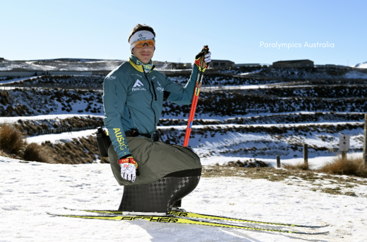 A sit-ski athlete in an Australian team jacket poses on snowy terrain, holding ski poles and wearing gloves, with mountainous slopes and buildings in the background. His adaptive sit-ski equipment is visible, and "Paralympics Australia" is written in white text. The image highlights para-athlete participation in winter sport.