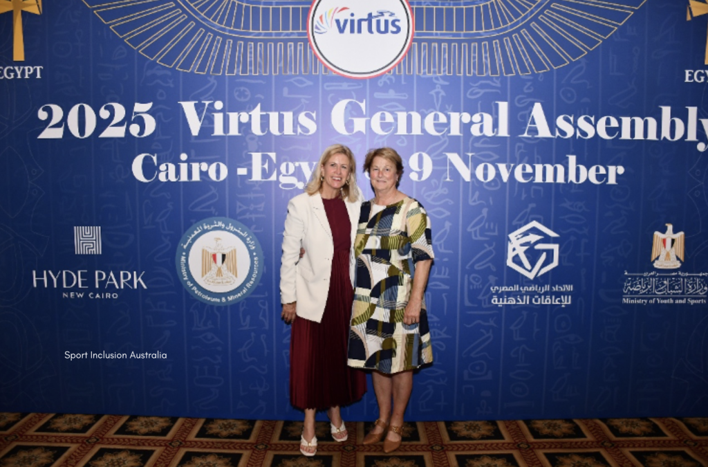 Two women stand together in front of a blue backdrop displaying the Virtus logo and text reading "2025 Virtus General Assembly, Cairo - Egypt, 9 November." The backdrop features gold accents and logos from sponsors and Egyptian government organisations. Both women are dressed in formal attire and are smiling for the photo, which appears to be taken at a professional event.