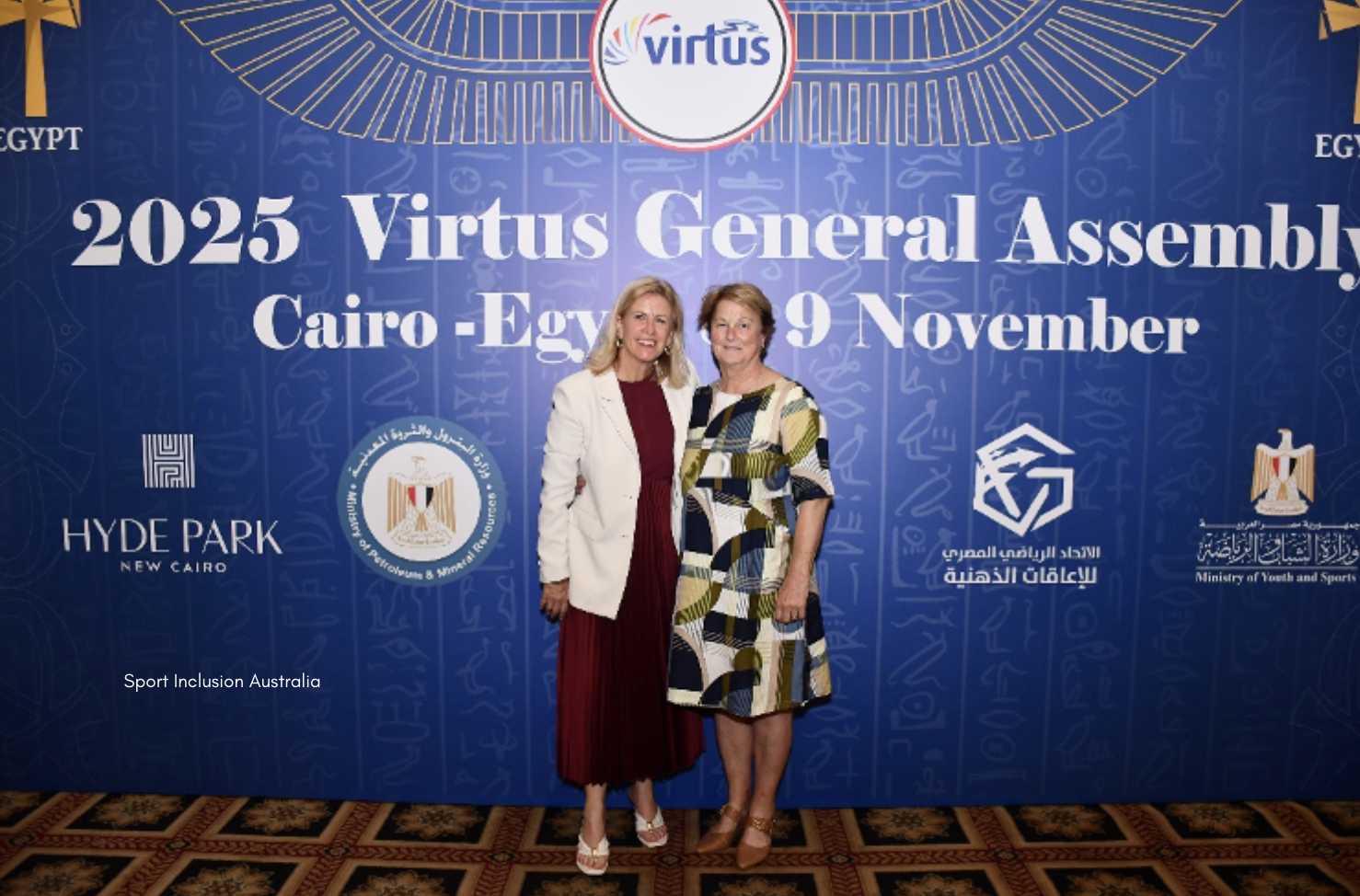 Two women stand together in front of a blue backdrop displaying the Virtus logo and text reading "2025 Virtus General Assembly, Cairo - Egypt, 9 November." The backdrop features gold accents and logos from sponsors and Egyptian government organisations. Both women are dressed in formal attire and are smiling for the photo, which appears to be taken at a professional event.