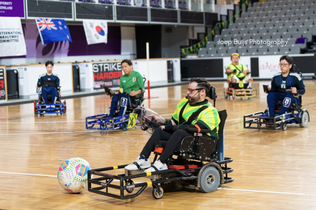 Several athletes compete in a game of powerchair football on a wooden indoor court; each player uses a powered wheelchair equipped with protective bumpers and distinctive-colored jerseys, maneuvering strategically around a large, multi-colored soccer ball. The court is surrounded by banners and flags, with empty stadium seating in the background, and the action is captured mid-play with a focus on teamwork and inclusivity.
