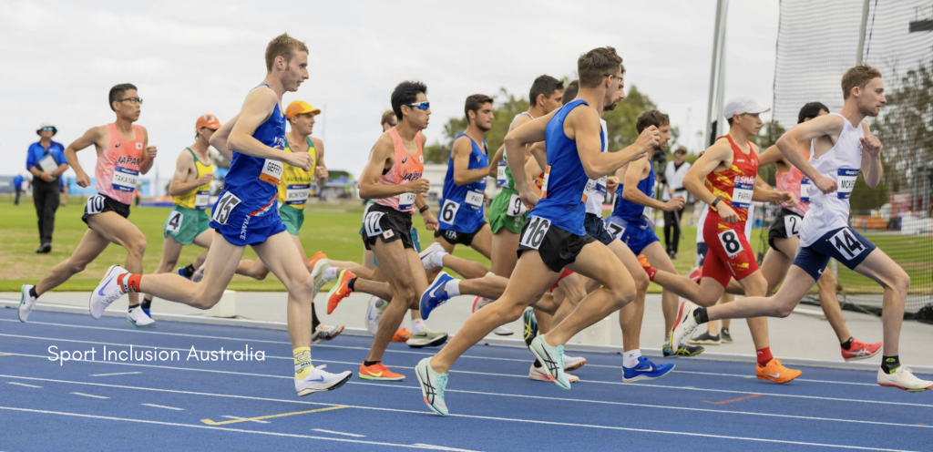 A group of male athletes with race bibs from different countries, including Australia, France, Japan, Italy, and Spain, are running on a blue track during an international athletics event. The runners are captured mid-stride, with focus and intensity as they compete. In the background, officials are watching and the grassy area of the track venue is visible. The image is captioned with "Sport Inclusion Australia."