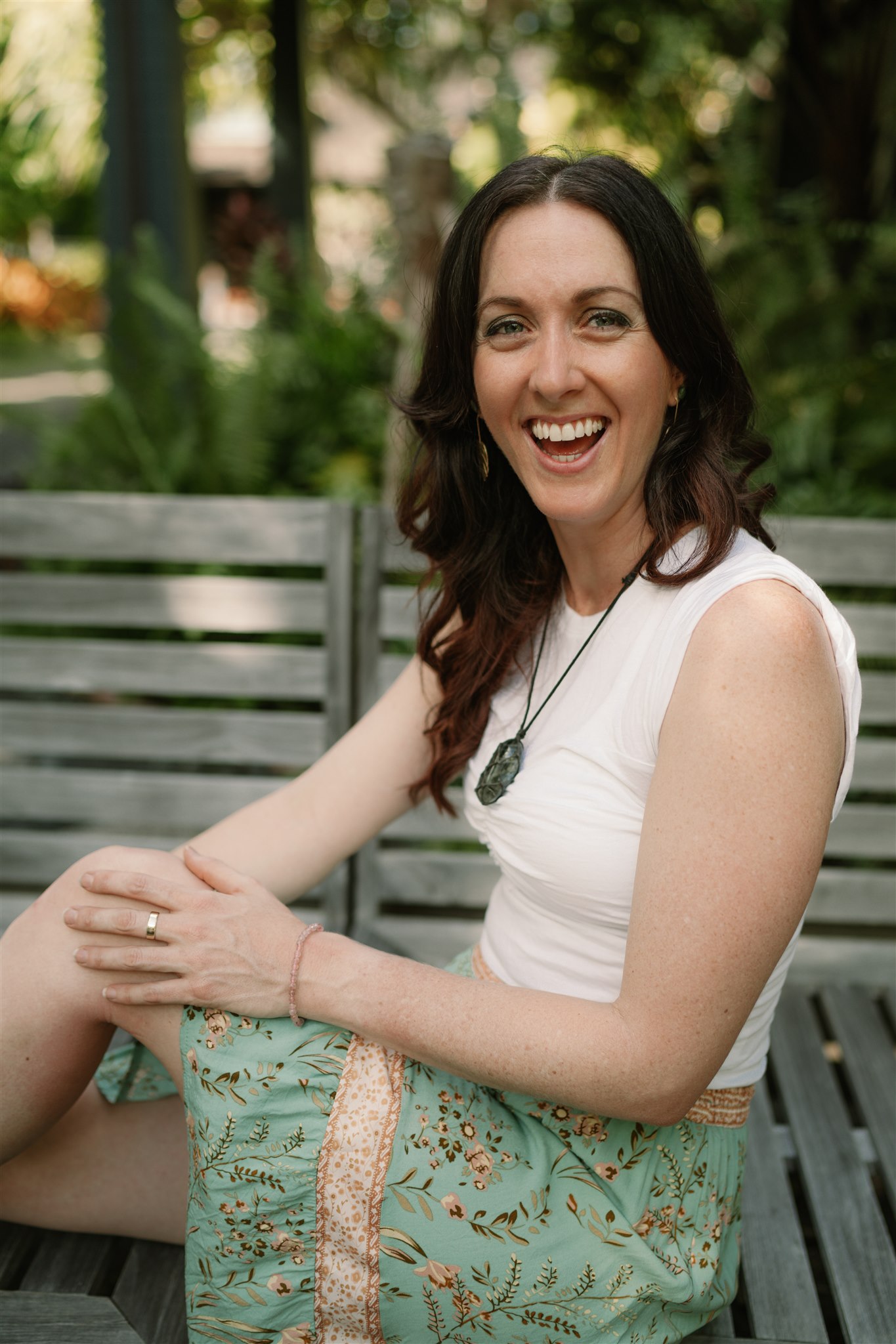 Jess poses with long wavy brown hair, wearing a white sleeveless top and a green floral skirt, sits on a wooden bench outdoors. She is smiling warmly at the camera, appearing relaxed and happy. Her hands rest on her knee, and she wears a dark pendant necklace and a gold ring. The background is lush and green with plants, suggesting a garden or park setting.