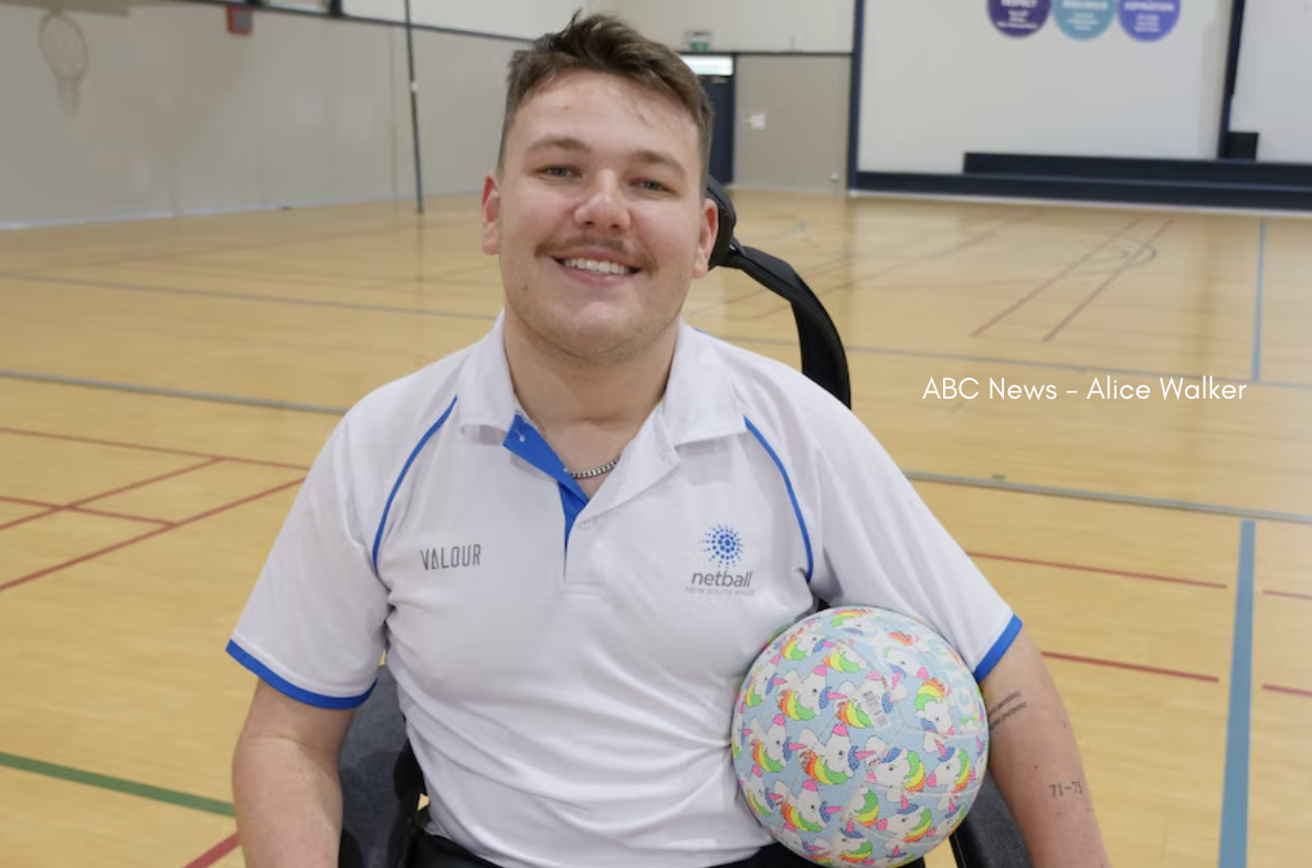 A wheelchair user wearing a white Netball New South Wales polo shirt sits on an indoor court holding a colourful unicorn-patterned netball, with basketball hoops and court lines visible in the background, and the photo credit “ABC News – Alice Walker” overlaid on the right side.