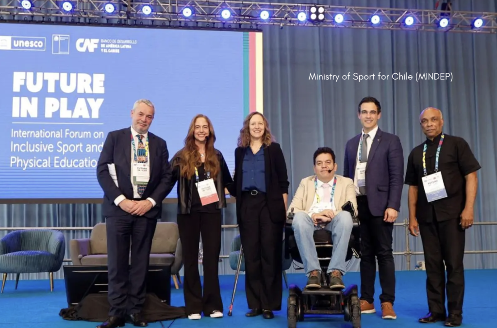 Six presenters stand side by side on a blue stage in front of a large screen reading “Future in Play – International Forum on Inclusive Sport and Physical Education,” with the UNESCO and CAF logos at the top; one person uses a power wheelchair, and all are wearing event lanyards under bright overhead stage lighting.