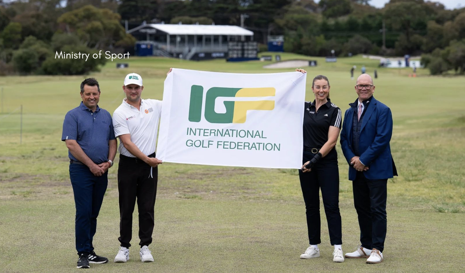 Four people stand on a golf course holding a large white International Golf Federation banner, with two people gripping the sides of the banner and two others standing on either end, and the text “Ministry of Sport” visible in the sky area on the left.