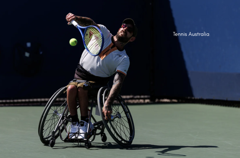 A wheelchair tennis player in a white shirt and cap leans forward on an outdoor hard court, extending a racquet to hit a yellow tennis ball, with strong sunlight casting shadows and a dark blue backdrop behind them.