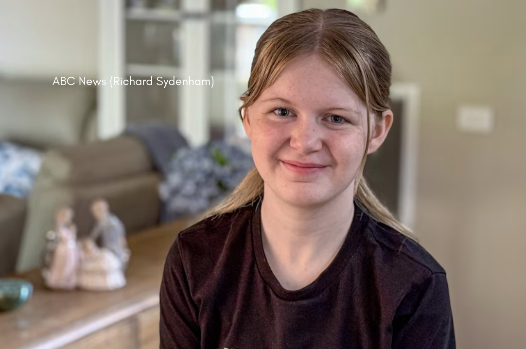 A smiling teenager with long light-brown hair tied back sits indoors wearing a dark T-shirt, with a softly focused lounge room and wooden table in the background.