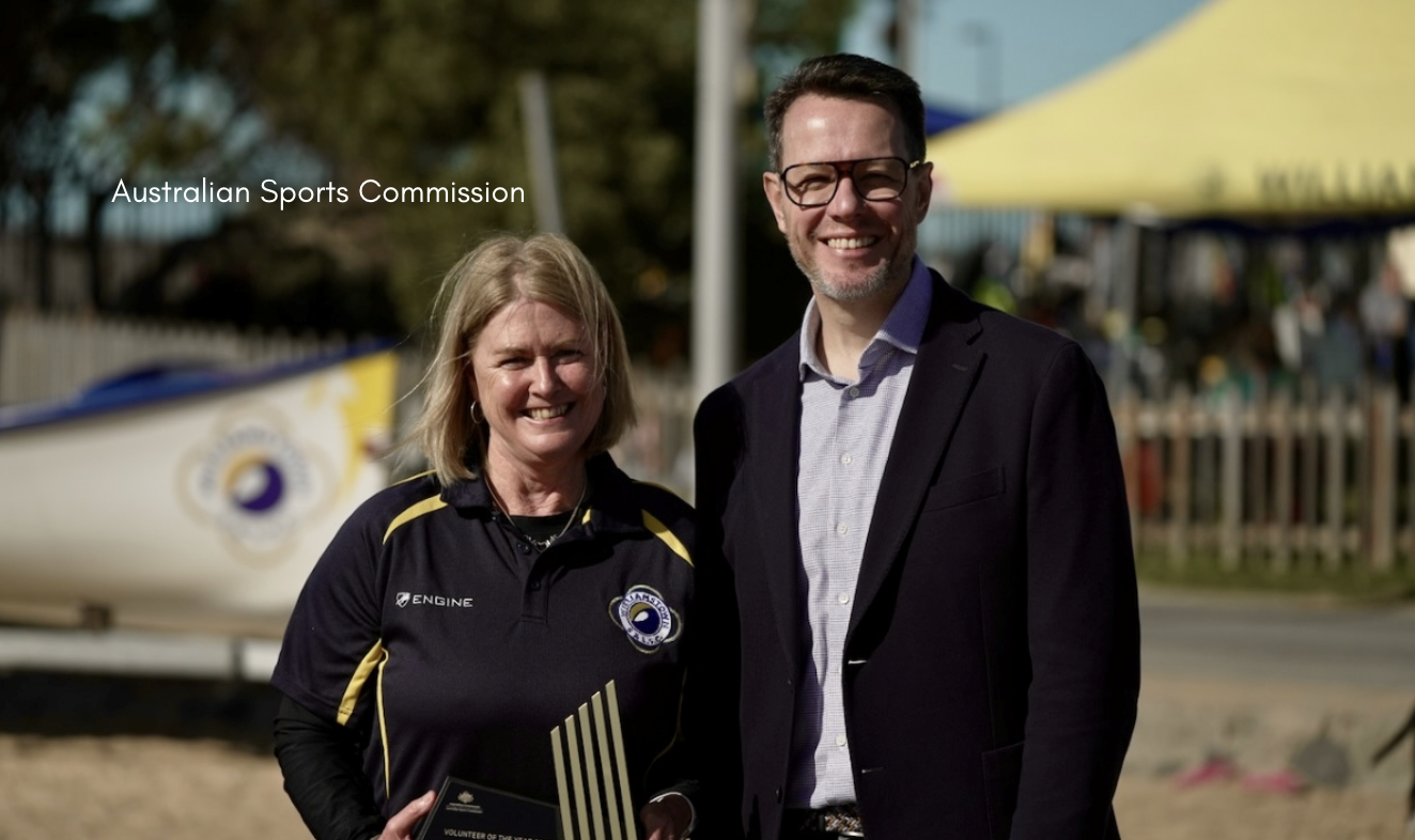 Two people stand side by side on the beach at Williamstown Life Saving Club, with the woman in a navy club shirt holding a Volunteer of the Year award and the man in a blazer beside her, as beach equipment and a yellow tent appear blurred in the background.