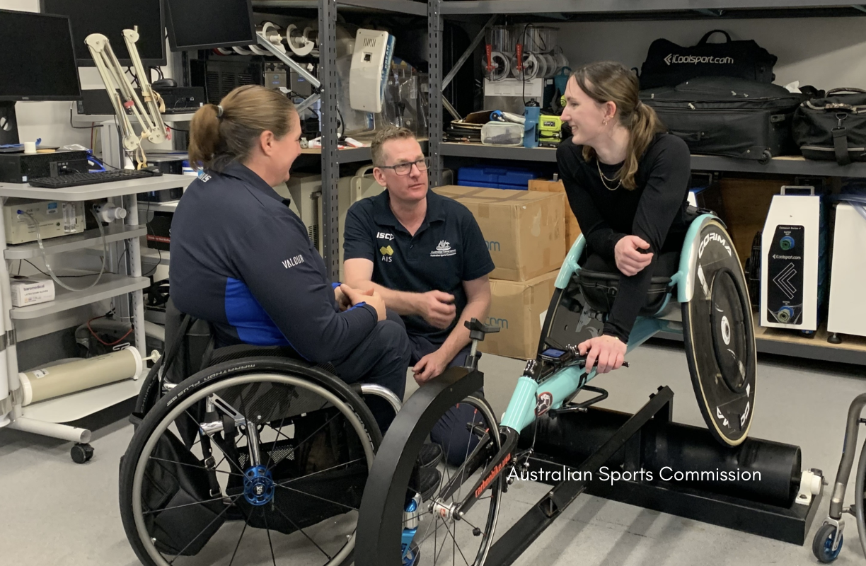 Two women wheelchair users and a man sit together in a sports engineering workshop, discussing and examining a teal racing wheelchair mounted on a roller, with shelves of equipment, tools and boxes in the background.