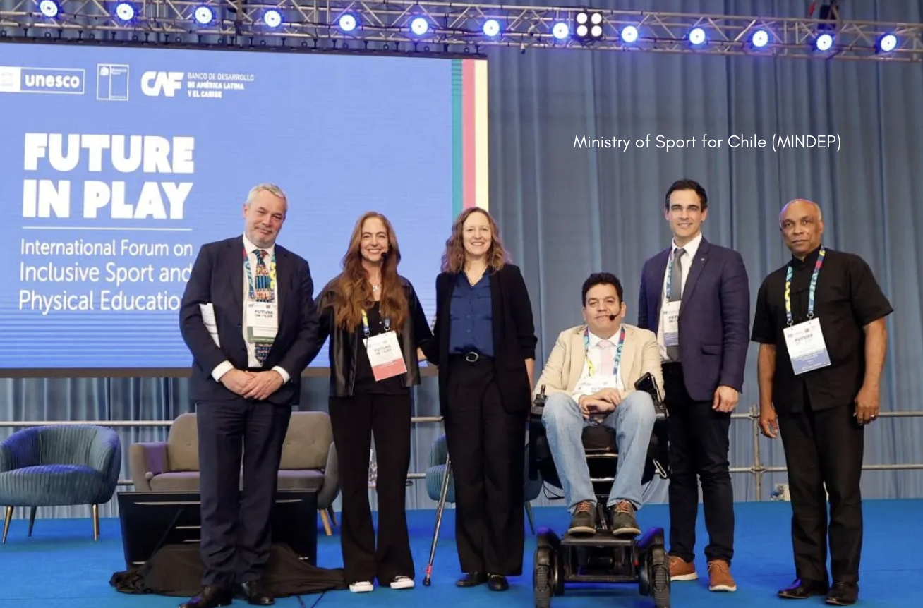 Six presenters stand side by side on a blue stage in front of a large screen reading “Future in Play – International Forum on Inclusive Sport and Physical Education,” with the UNESCO and CAF logos at the top; one person uses a power wheelchair, and all are wearing event lanyards under bright overhead stage lighting.