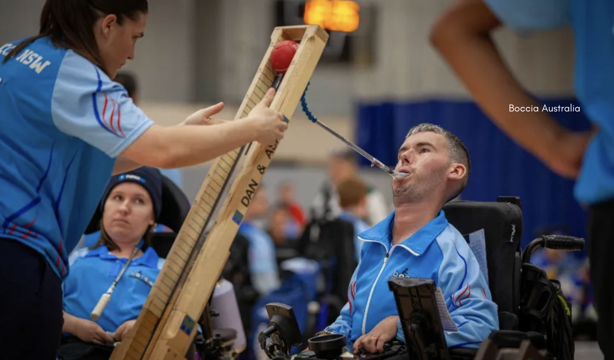 A boccia athlete in a powerchair lines up a shot using a head pointer and ramp, while a support person in a NSW team shirt steadies the ramp and another athlete looks on in the background at an indoor boccia competition.