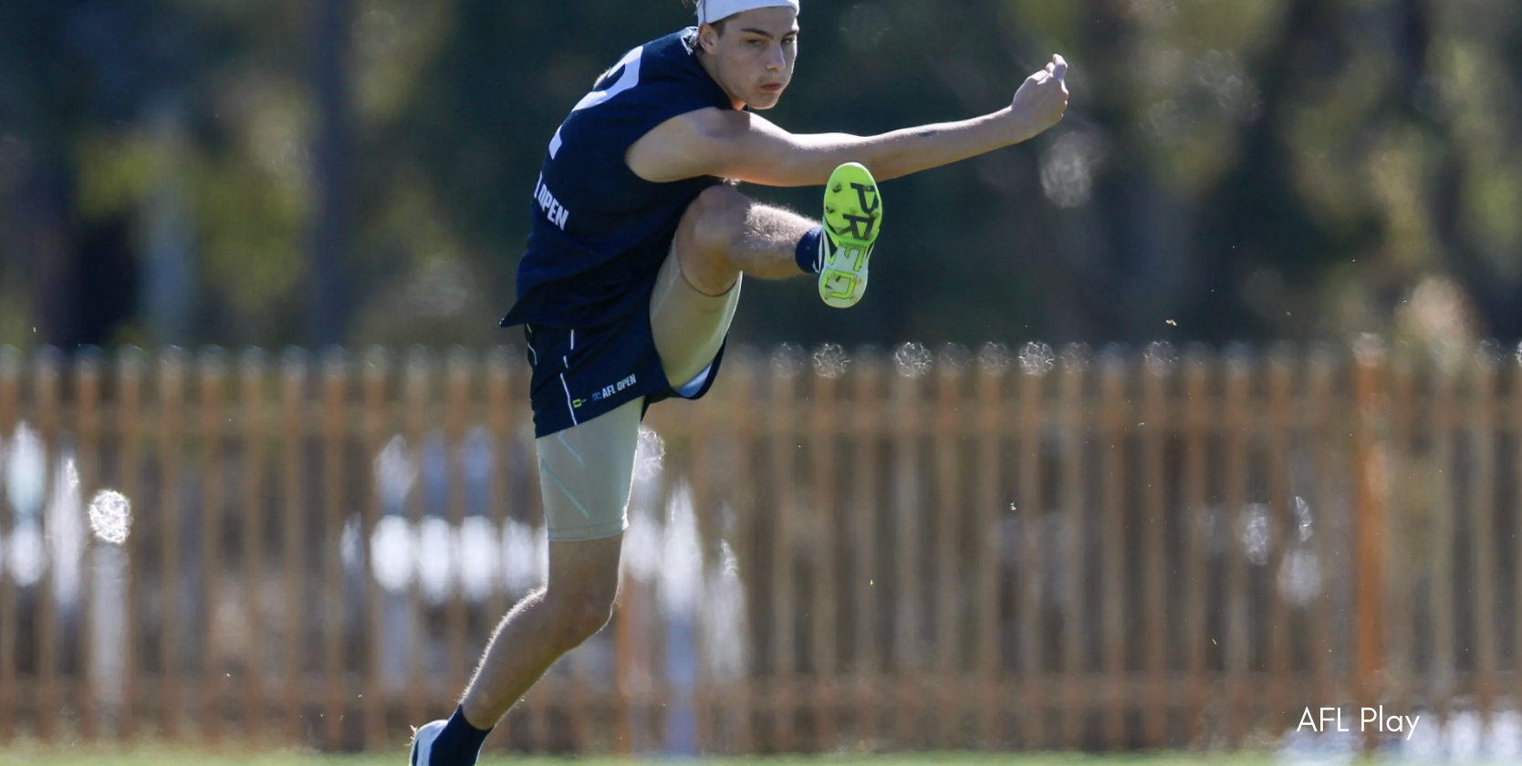 A footballer with a blurred face kicks the ball mid‑air on a sunny oval, with one leg extended high and a wooden boundary fence and trees softly out of focus in the background.
