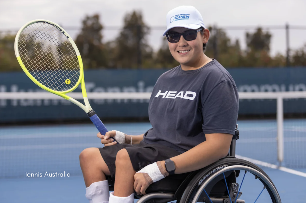 A wheelchair tennis player sits on an outdoor hard court holding a tennis racquet, wearing a Melbourne Open cap, HEAD T-shirt and sports watch, with the rear wheel of the sports wheelchair visible in the foreground.