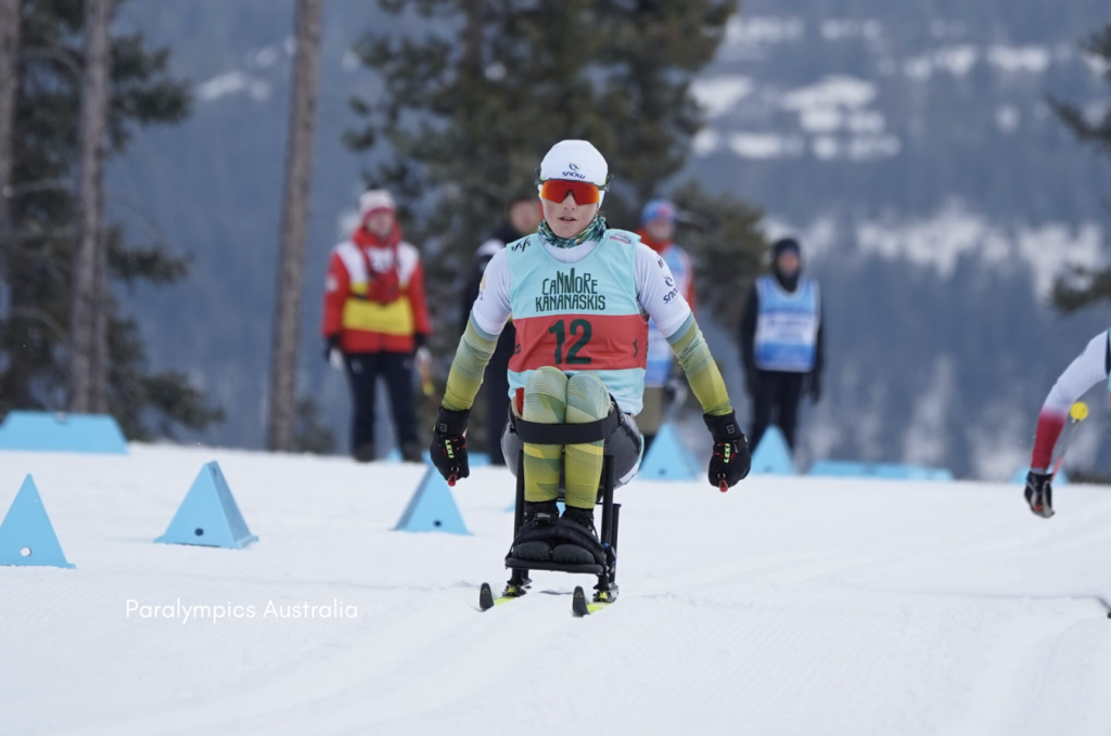 A Para Nordic sit-skier wearing Australian race colours and a bib marked “12 Canmore Kananaskis” pushes along a snowy course, with officials and other competitors blurred in the background and snow-covered trees behind them.