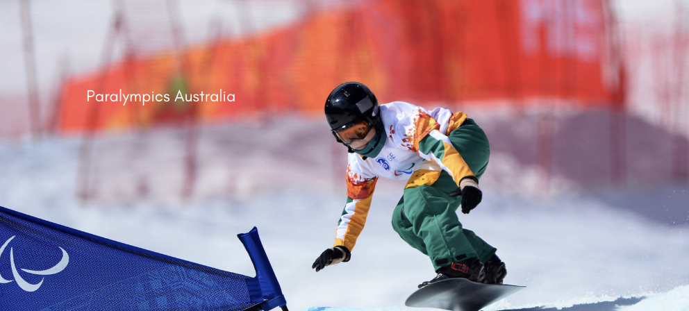 A Para snowboarder in an Australian team uniform races down a snowy course, knees bent and leaning forward on a snowboard, with a blue Paralympic gate flag in the foreground and blurred red fencing in the background.