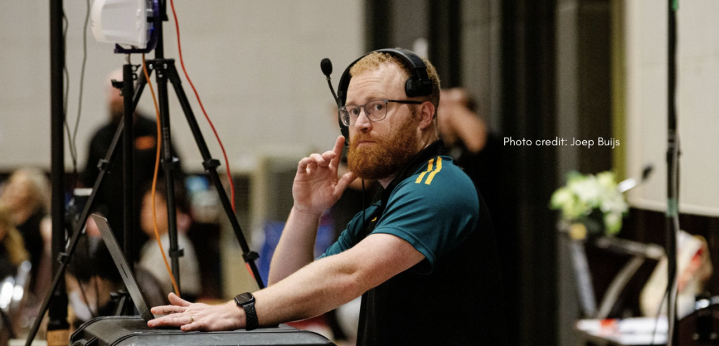 Man wearing a green and gold polo shirt and headset stands at a courtside technical table, one hand on equipment and the other near his face as he focuses on the game action in an indoor sports venue.