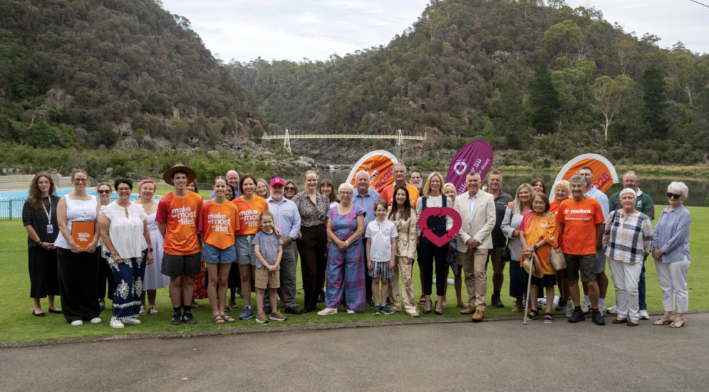 A large group of people, including adults and children, stand together on a grassed area in front of a river and tree-covered hills at Cataract Gorge in Launceston. Many are wearing bright orange T-shirts featuring transplant and donation messaging, while branded event banners are visible behind them, suggesting a community launch or announcement related to the Australian Transplant Games.