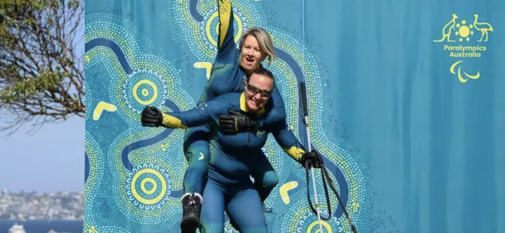 Two para‑skiers in matching teal and yellow skinsuits perform a tandem pose in front of a bright turquoise Paralympics Australia backdrop featuring Indigenous‑inspired circular and flowing dot patterns, with one athlete lifting the other piggyback style and raising an arm triumphantly, ski equipment visible at their side and a blurred coastal landscape in the far background.