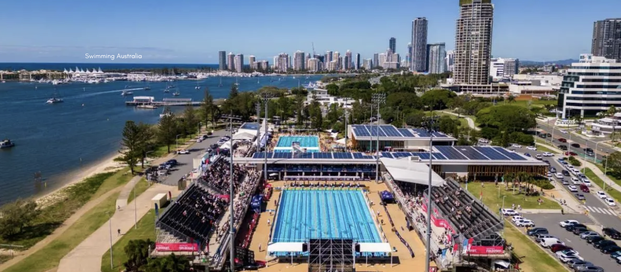 Aerial view of an outdoor swimming complex on the waterfront, with a 50‑metre pool in the foreground surrounded by grandstands full of spectators, additional pools and buildings behind, a riverside path and parked cars to the left, city high‑rise skyline in the background under a clear blue sky, and the waterway dotted with boats on the left side of the image.