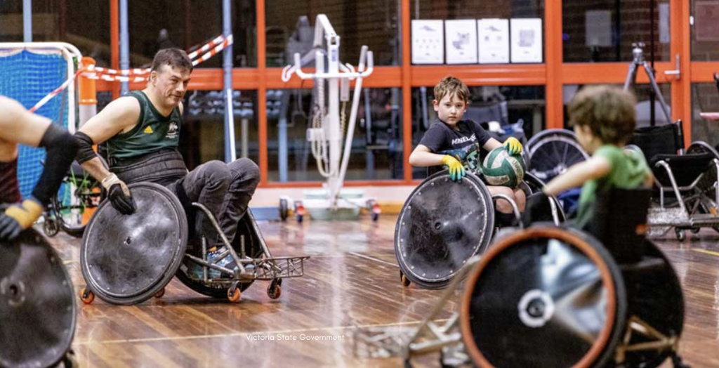 Indoor wheelchair rugby (or wheelchair basketball) training session on a polished wooden court, showing several athletes in sport wheelchairs, one holding a ball, moving and facing each other in play, with sports equipment, spare wheelchairs, and large windows and doors in the background.
