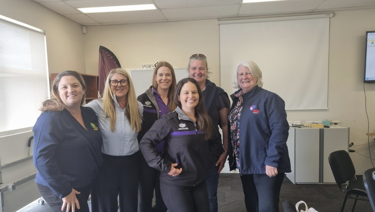Group of six women from different sporting and disability organisations standing together and smiling in a meeting room, wearing branded uniforms and jackets, with a projector screen and office furniture in the background.