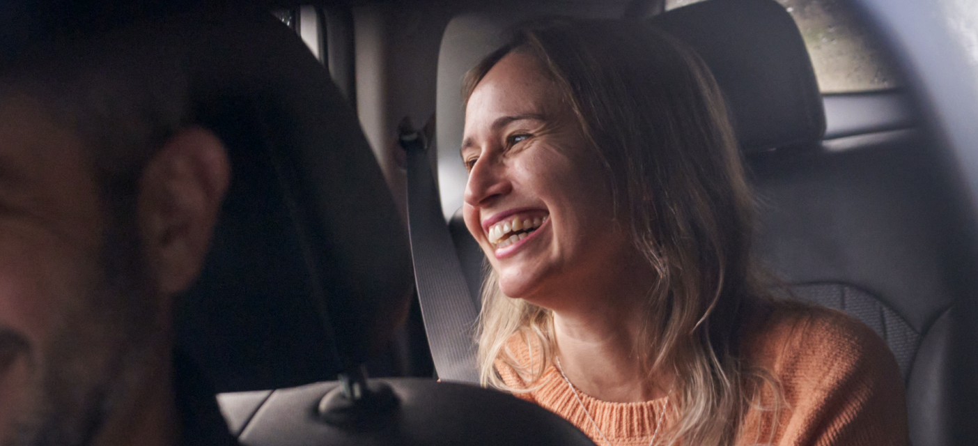 A person wearing an orange knit sweater sits in the front passenger seat of a car, with their face blurred for privacy, while another person is partially visible driving; both are wearing seatbelts and the interior of the vehicle is dimly lit.