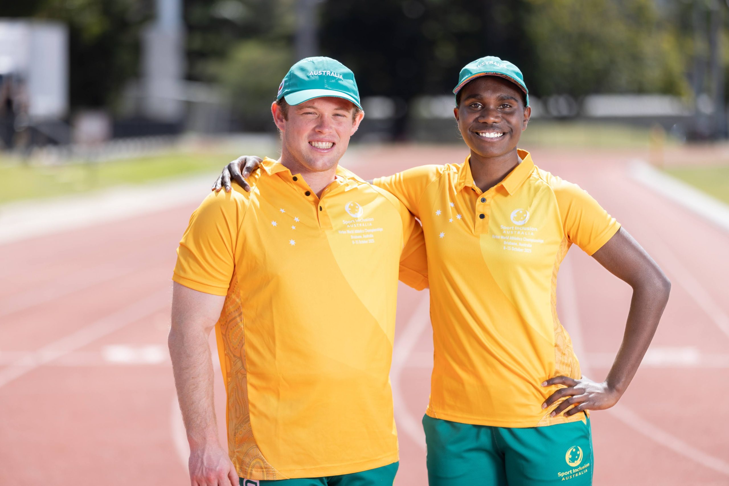 Two track and field athletes in yellow Sport Inclusion Australia shirts and green shorts stand arm in arm on the running track, smiling toward the camera.