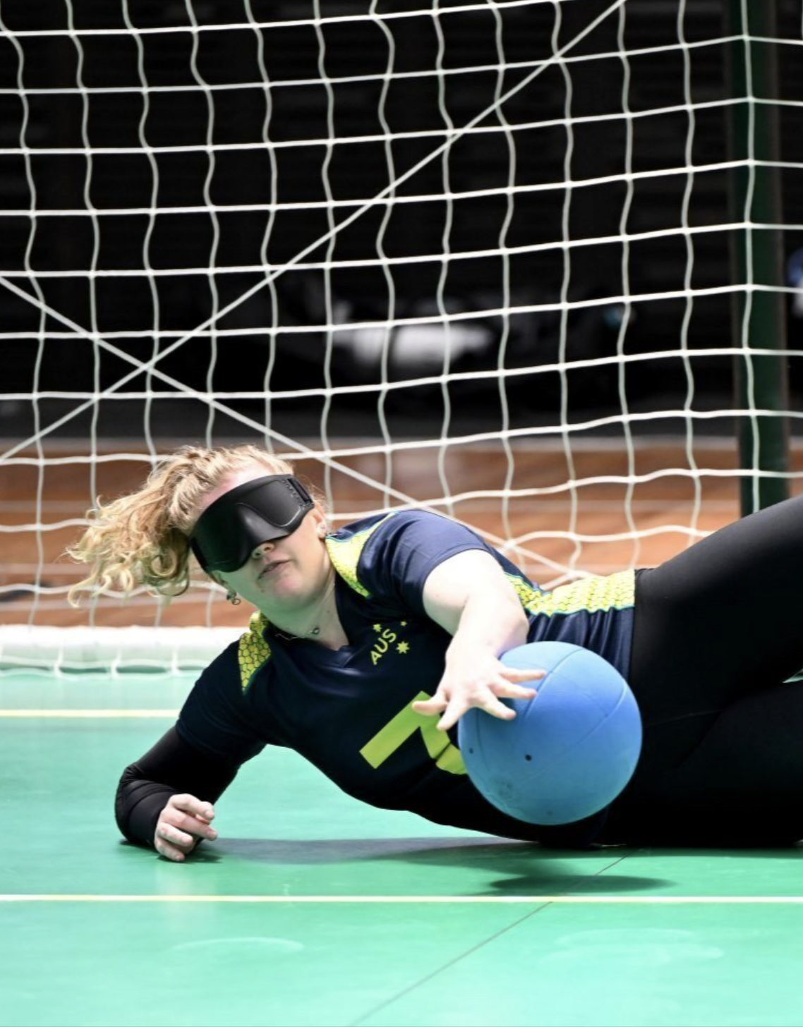 Goalball player Brodie Benson dives across the court in an Australian uniform, blocking a blue ball in front of the net.