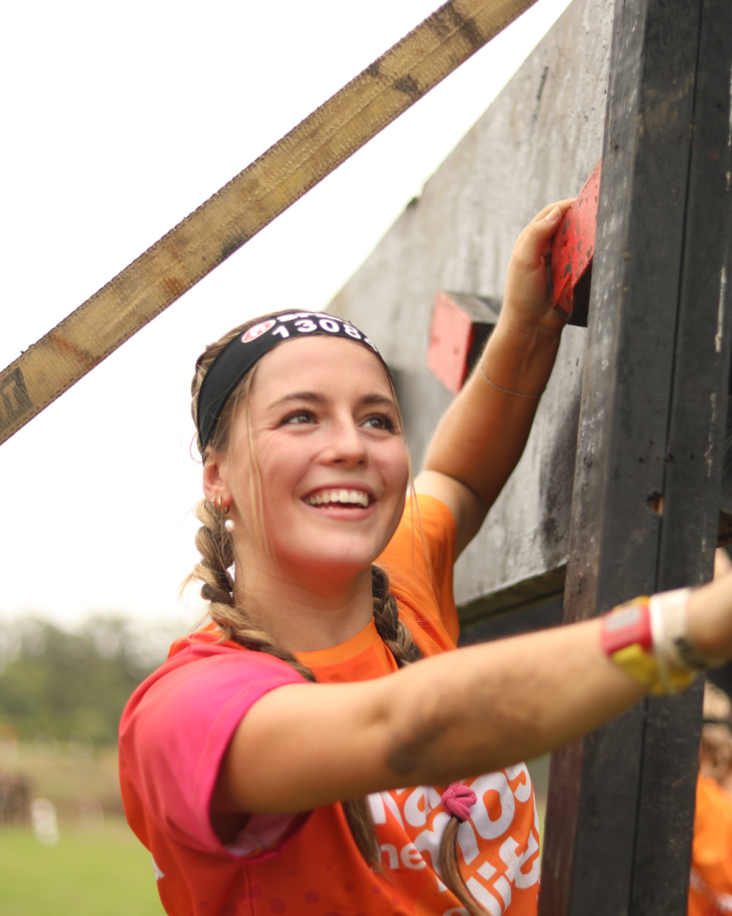 Young woman in a bright orange Transplant Australia shirt and headband climbs over a wooden obstacle, gripping the top edge with muddy arms during an outdoor course.