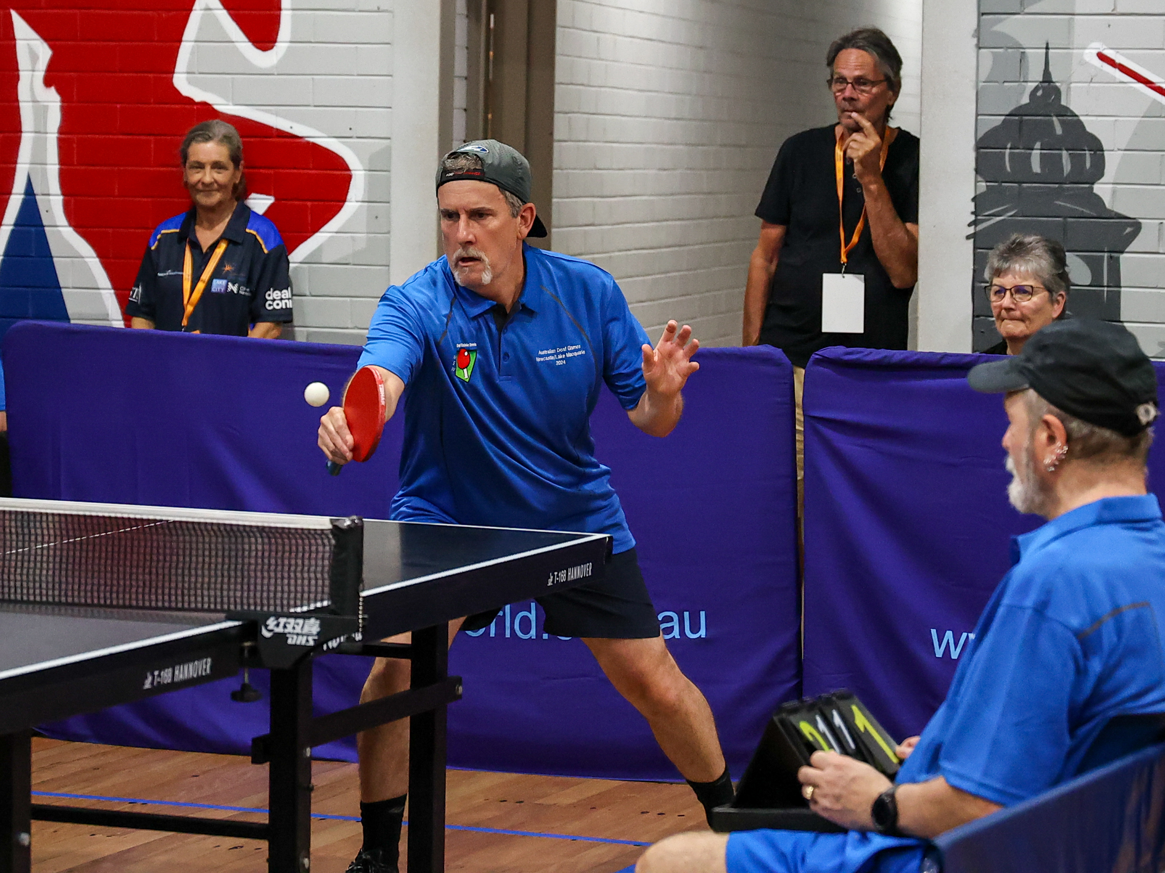 Table tennis player in a blue team shirt serves the ball during an indoor match, watched by an official and seated scorer in matching blue uniforms.