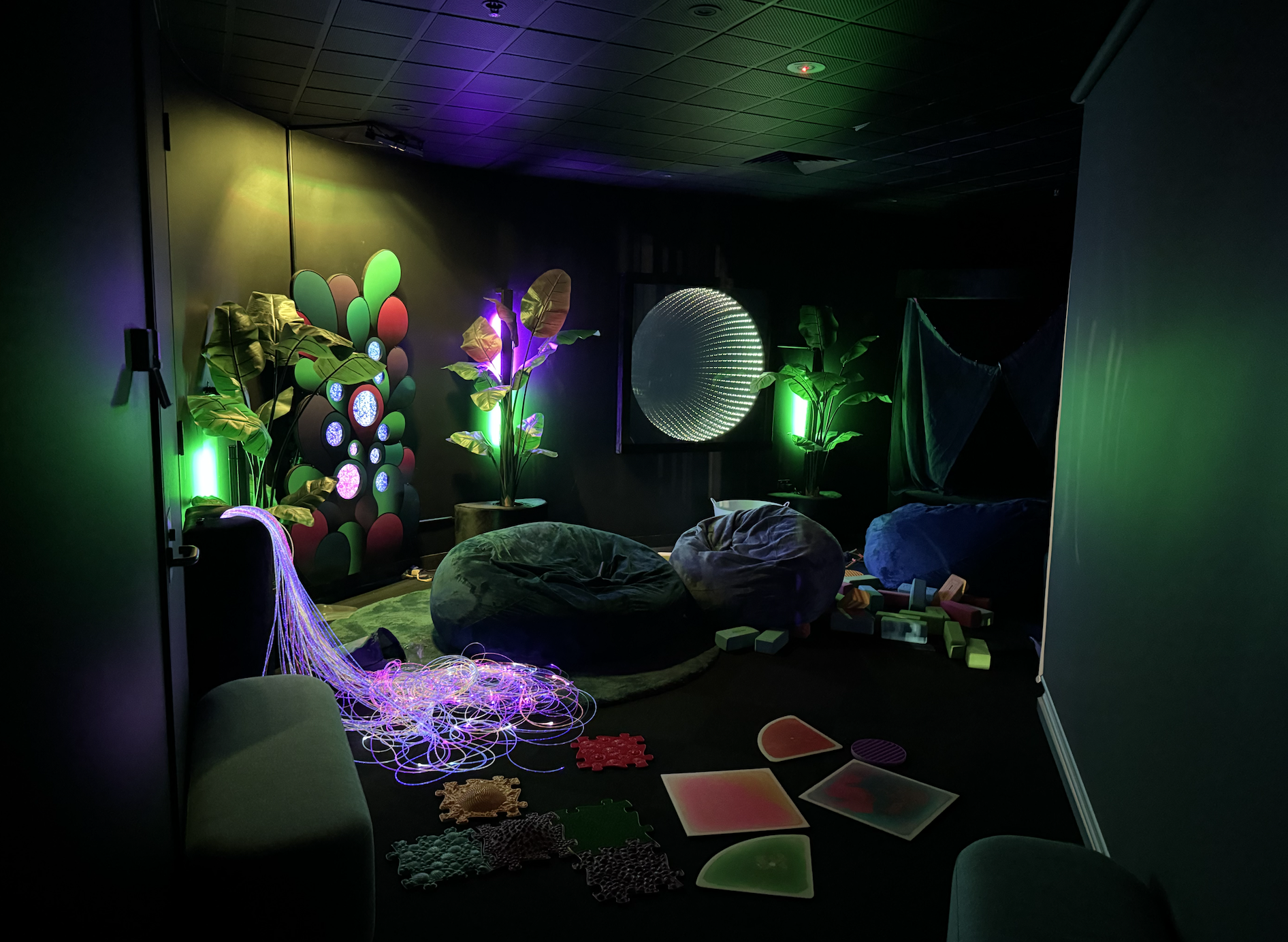 A dark, calming sensory room with beanbags, soft mats and scattered tactile tiles on the floor, surrounded by glowing green and purple lights, plants and interactive wall panels creating a quiet, immersive space.
