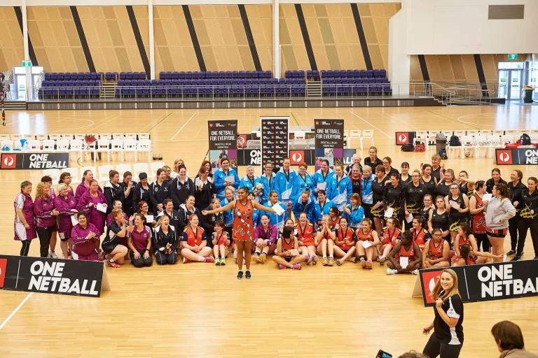 Netball players and team staff gather for a group photo on an indoor court at the 2017 Marie Little Shield. Wearing colourful team uniforms, they sit and stand together, with many smiling and applauding.