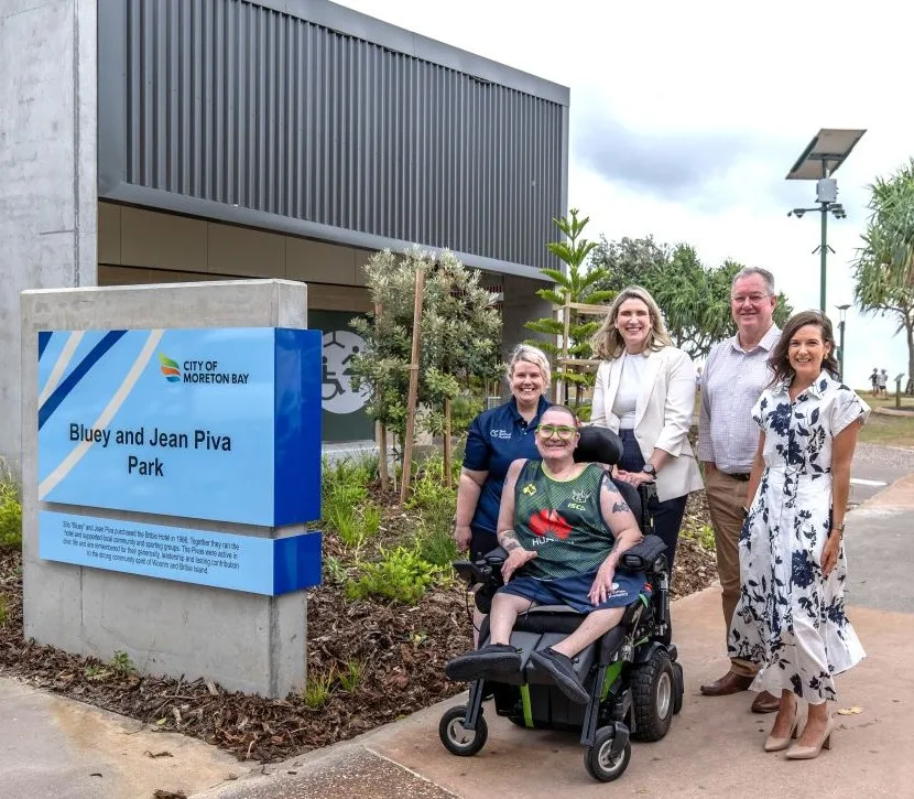 Five people, including a person seated in a powered wheelchair, smile for a photo outside the new accessible amenities building at Woorim Beach. Beside them is a blue City of Moreton Bay sign reading “Bluey and Jean Piva Park”, surrounded by landscaped gardens.