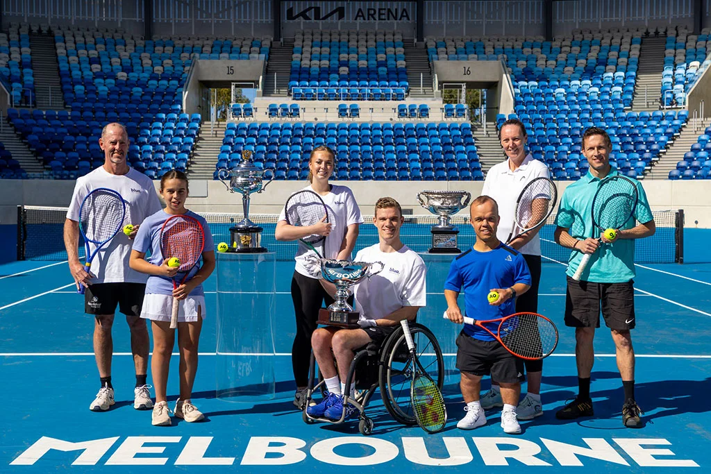 Seven people, including a wheelchair user, pose on a blue tennis court at Kia Arena in Melbourne. They hold tennis racquets and balls, with three trophies displayed among the group. Rows of blue stadium seats fill the background.