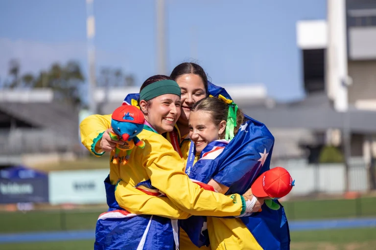 Emma Papantoniou, Siarne Davis and Claudia Flamini embrace and smile at the 2025 Virtus World Athletics Championships in Brisbane, wearing Australian team uniforms and draped in Australian flags while holding event mascots.